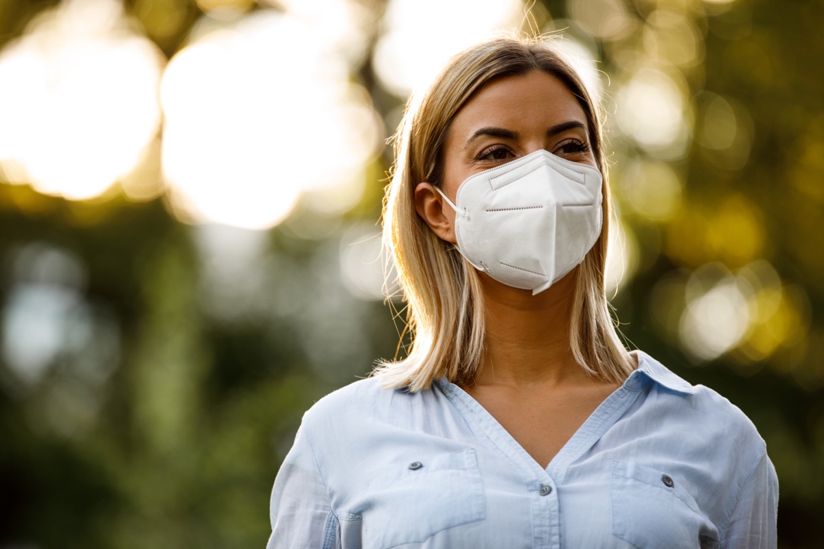 Young woman standing at the public park, enjoying a beautiful day out. She is smiling behind N95 face mask she is wearing.