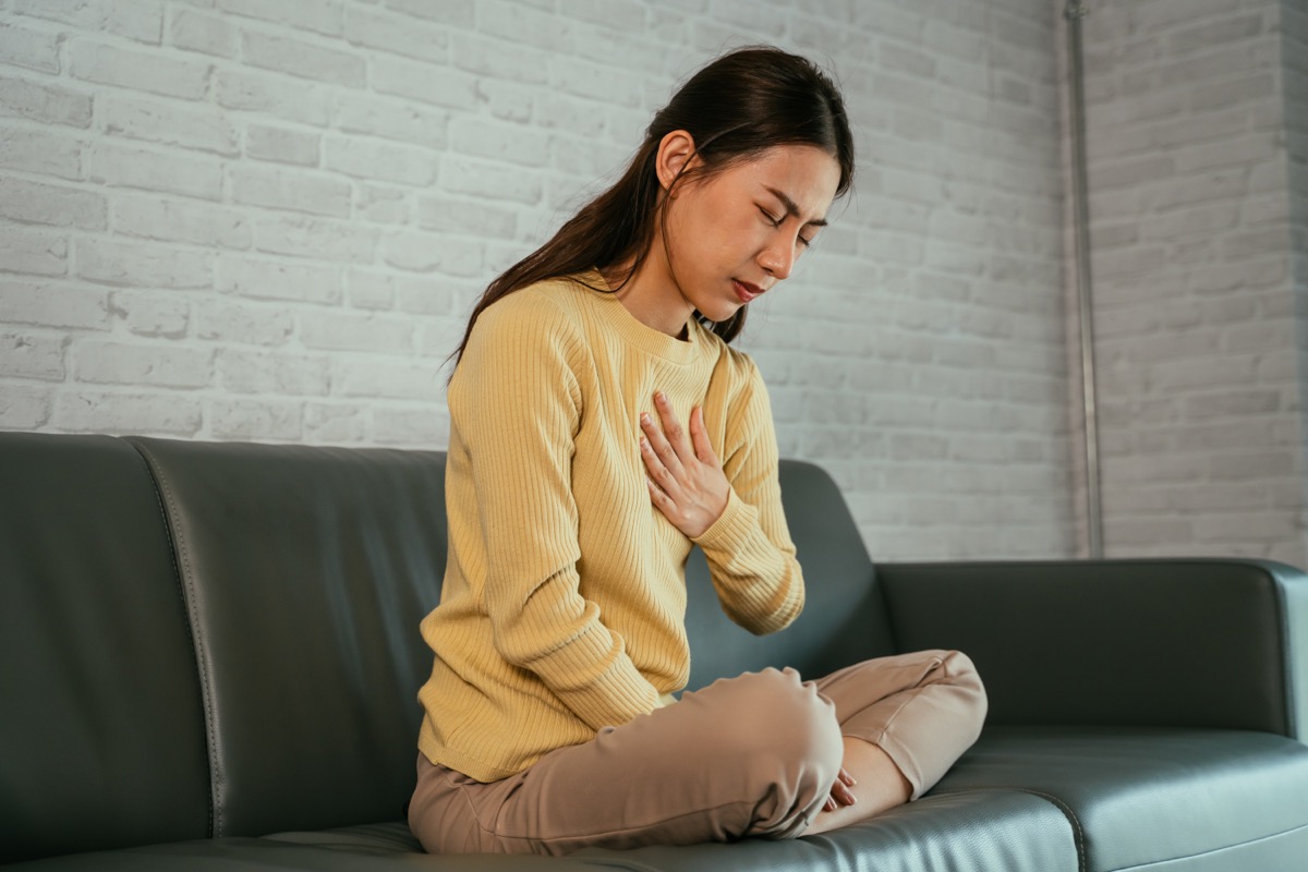 Asian young woman feeling discomfort as suffering from heartburn holding chest with closed eyes and sitting with folded legs on couch at home.