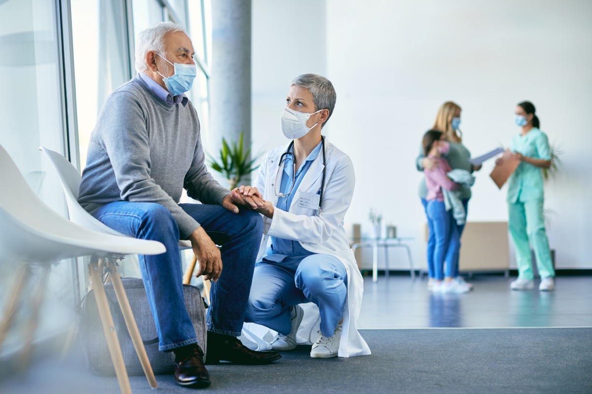 Distraught senior man sitting at hospital waiting room while female doctor is holding his hand and comforting him