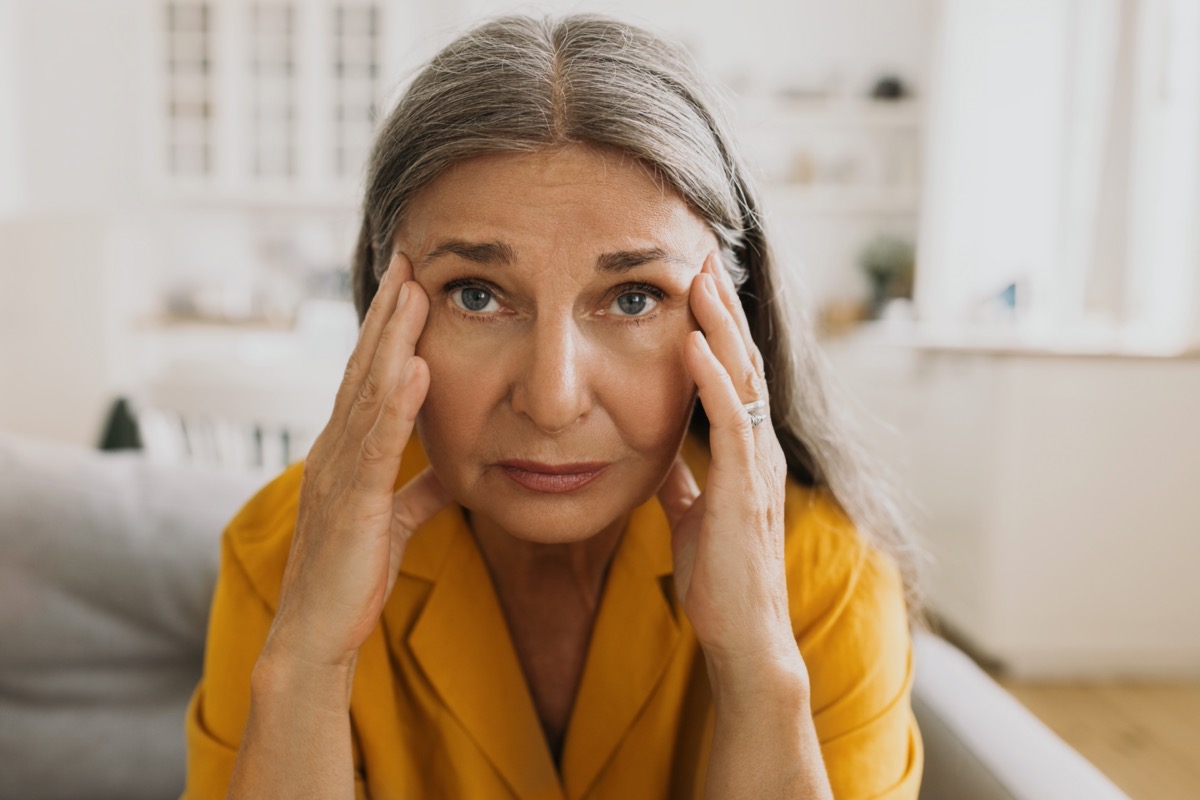 A woman suffering from stress and headache, sitting on couch massaging temples with fingers, looking at camera with painful face expression.