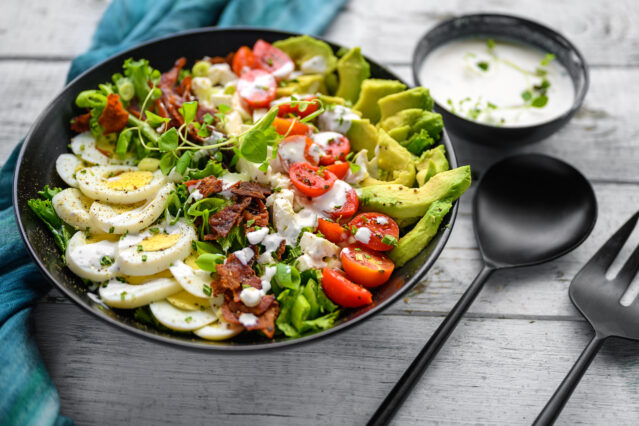 Cobb salad in black bowl with dressing on the side, black serving spoons, blue napkin.