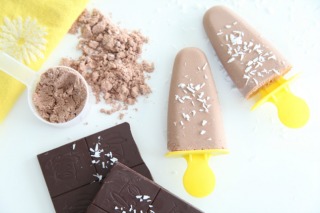 Two fudge pops with yellow popsicle sticks next to squares of dark chocolate and chocolate collagen powder.