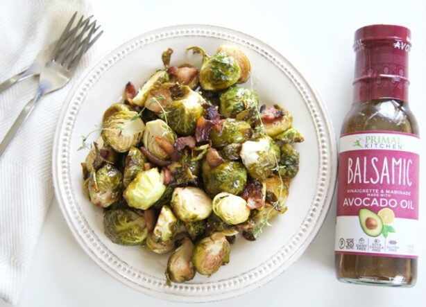 Plate of roasted Brussels sprouts and bacon alongside a fork and a bottle of Primal Kitchen Balsamic Vinaigrette.