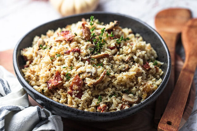 Black bowl filled with cauliflower bacon mushroom risotto. Wooden serving spoon.