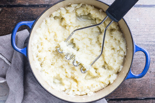 Mashing cauliflower in a blue enamel pot with a potato masher. 