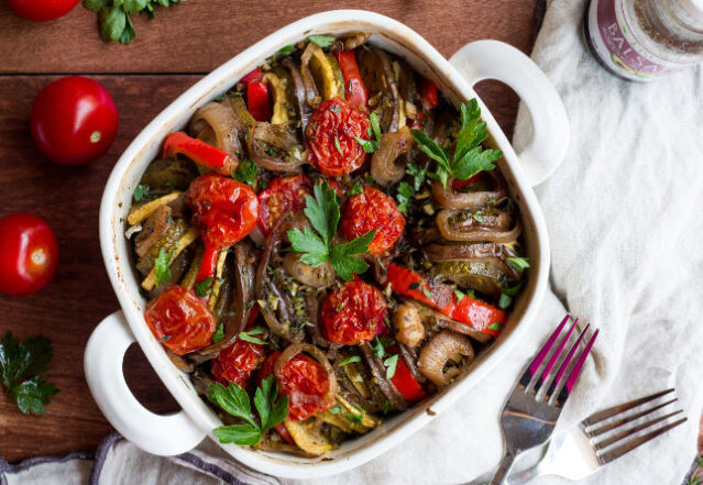 White casserole dish of ratatouille on a wooden background with white towel and fork.