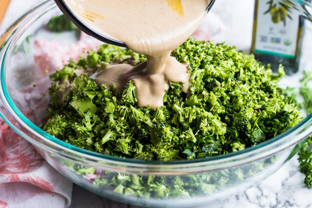 Pouring tahini dressing over finely chopped broccoli salad in a glass bowl.