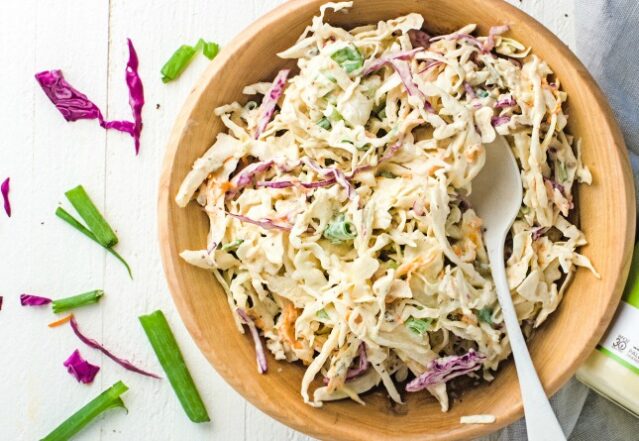 Cole slaw in a yellow ceramic bowl with a silver spoon on a white background adorned with green onions and purple cabbage.