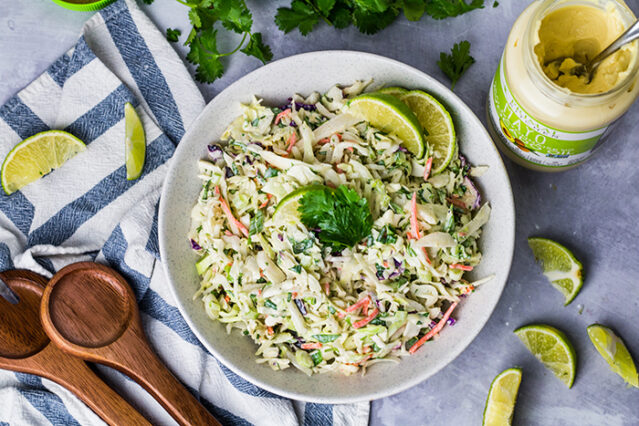 Overhead shot citrus cole slaw in a white bowl with wooden spoons, sliced limes, and a jar of Primal Kitchen Mayo.