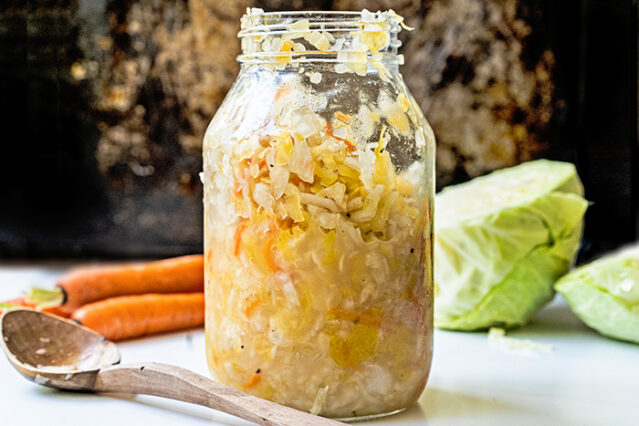 Large, open jar of sauerkraut in front of a head of <a href=