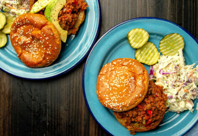 Sloppy joes sandwiches on blue plates with pickles and a side of cole slaw.