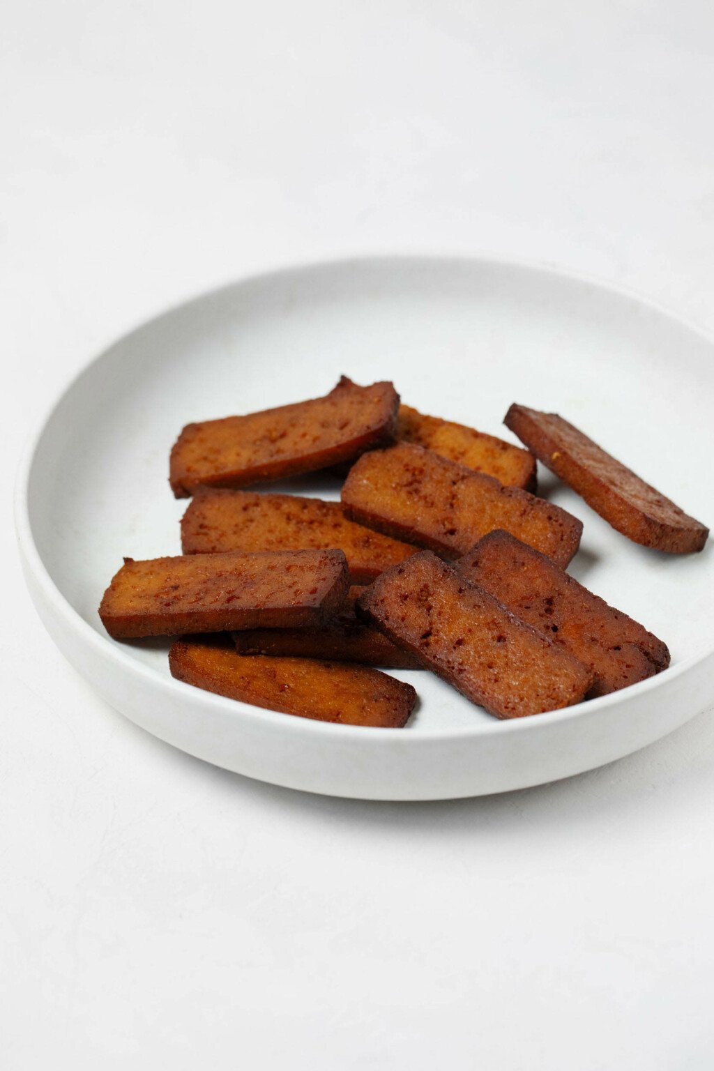 A round, low white bowl is filled with chewy slices of marinated, baked bean curd.