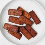 A round, white plate has been covered with rectangular, dark brown slices of baked, smoky tofu.