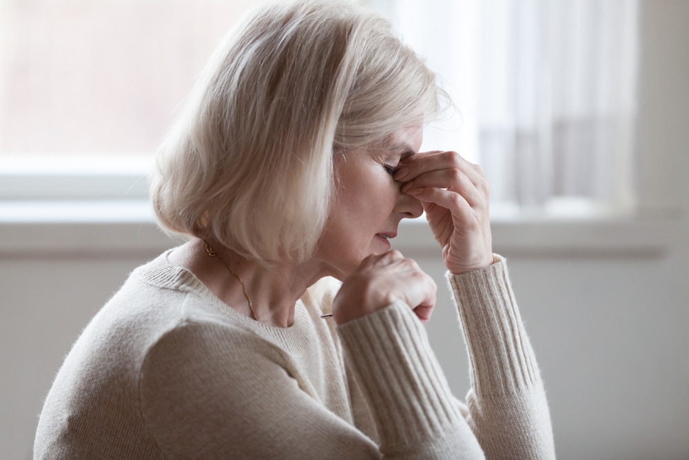 Distressed older woman with her eyes closed pinching the bridge of her nose to relieve a headache.