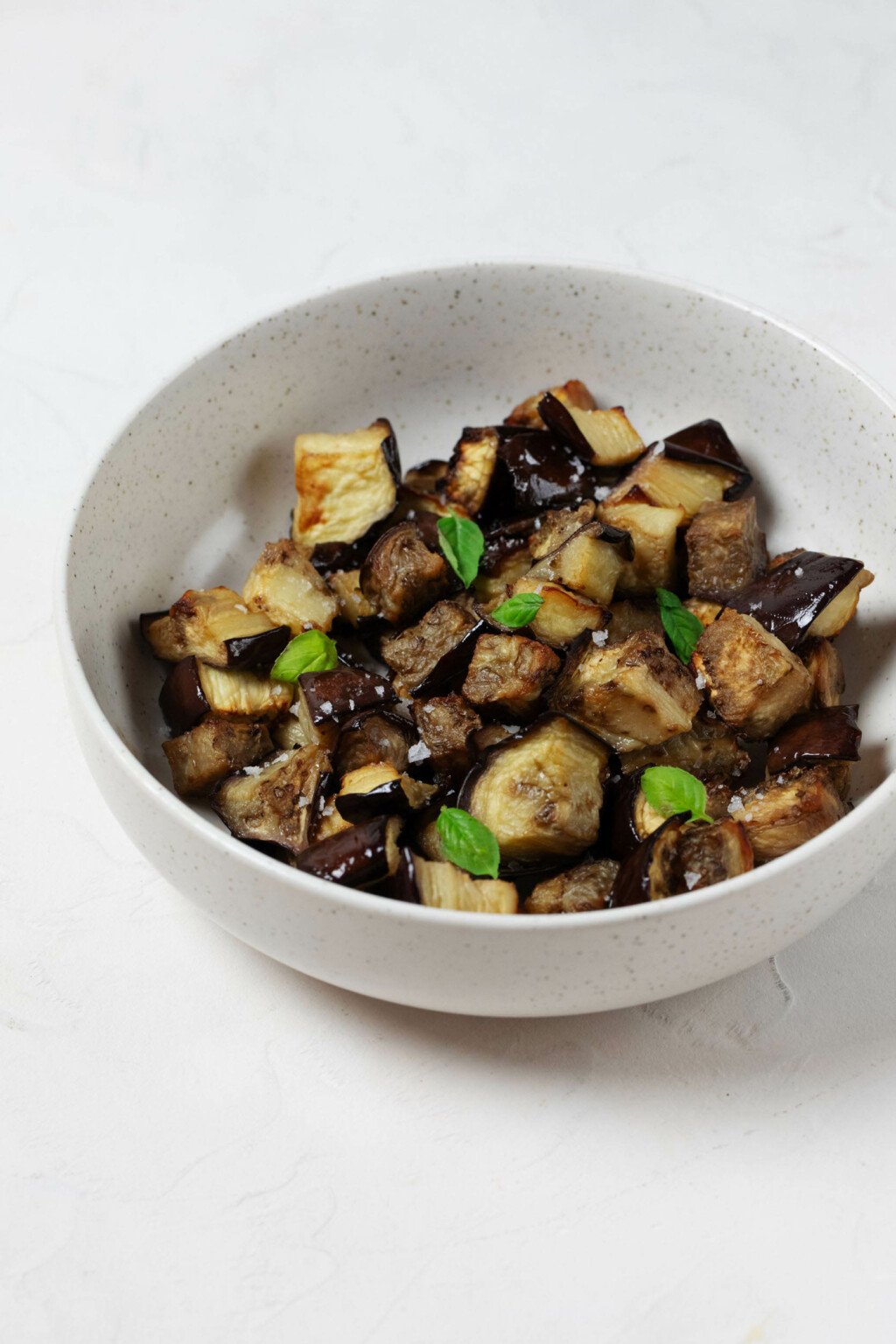 A white, ceramic bowl is filled with oven roasted eggplant cubes and tiny leaves of fresh, green basil.