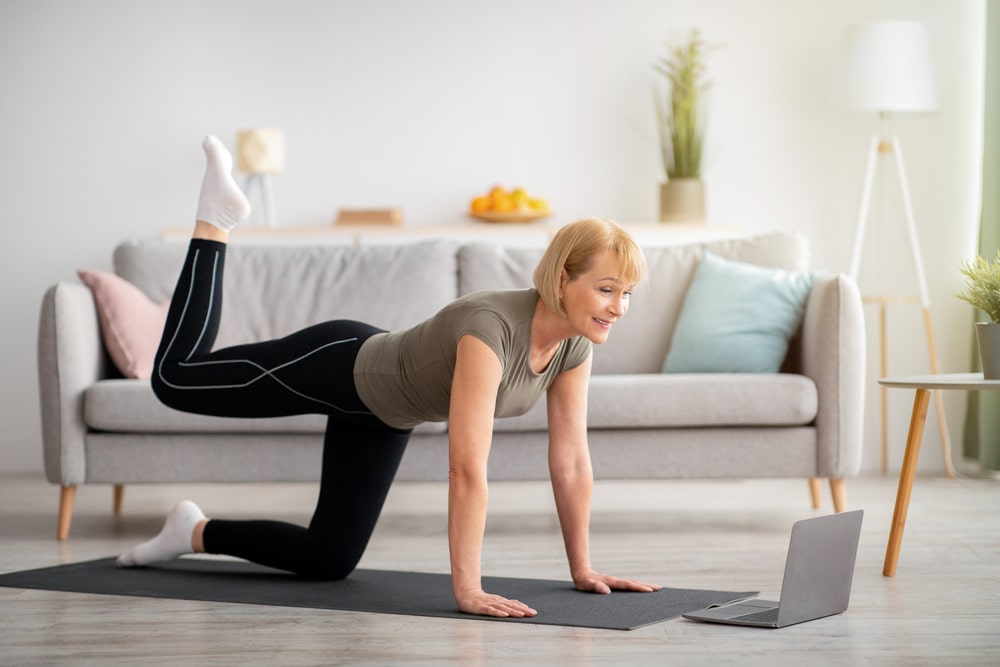 An older woman doing leg lifts on a yoga mat in front of her laptop.