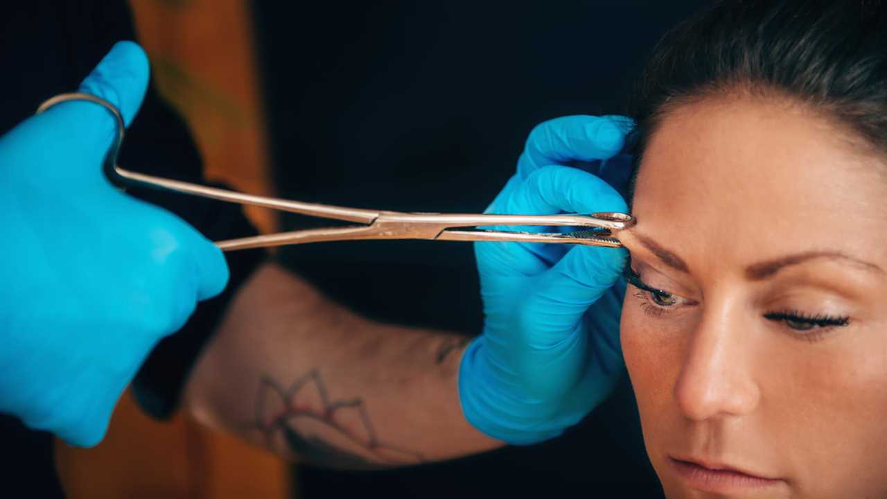 Beautiful young woman getting her eyebrow pierced, body piercer wearing blue gloves