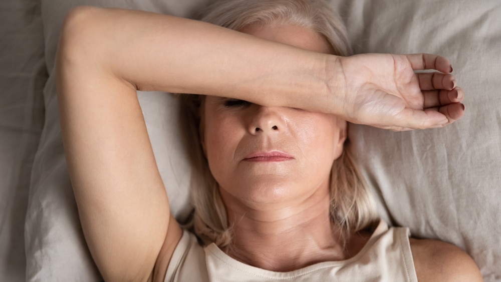 Close up top view of a middle-aged woman lying down in bed on a pillow putting her arm over her face.