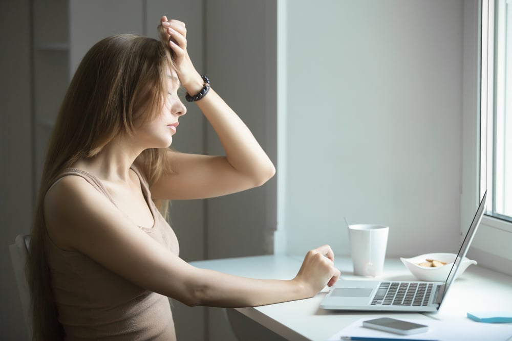 A tired woman sitting at a table with her laptop putting her hand on her head.